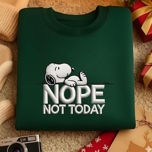 May include: A dark green sweatshirt featuring an embroidered Snoopy graphic lying on grass with the words "NOPE NOT TODAY" in white. The sweatshirt has a crew neck and is made of a soft material. The design is centered on the front of the garment.