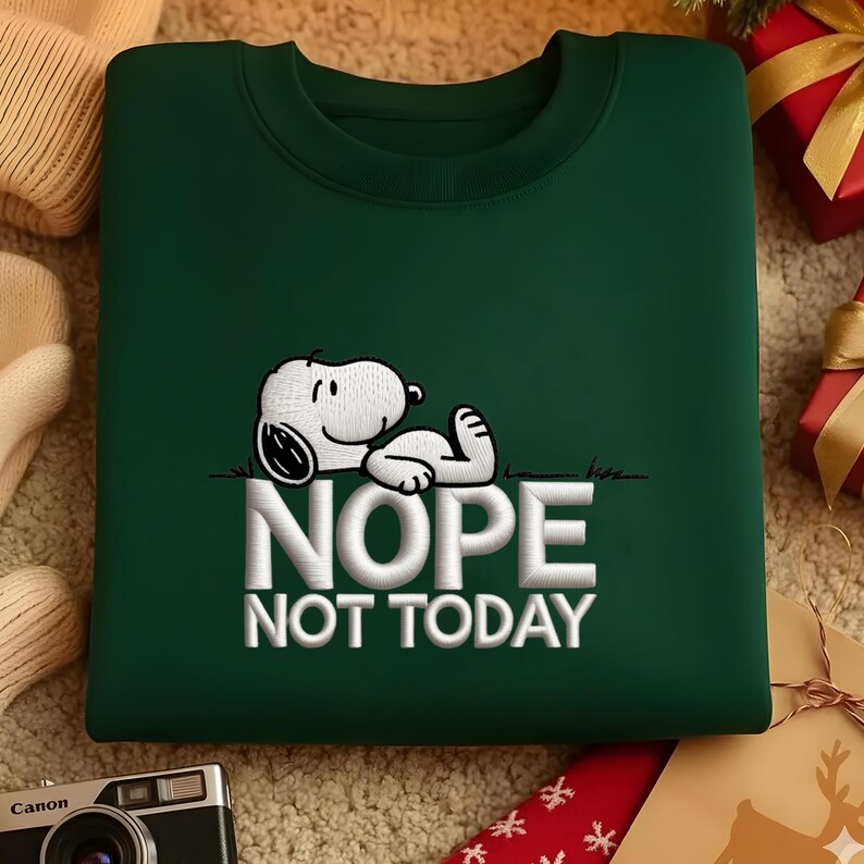 May include: A dark green sweatshirt featuring an embroidered Snoopy graphic lying on grass with the words "NOPE NOT TODAY" in white. The sweatshirt has a crew neck and is made of a soft material. The design is centered on the front of the garment.