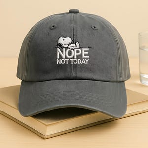 May include: A gray baseball cap featuring an embroidered Snoopy design with the words "NOPE NOT TODAY" in white. The cap has a curved brim and a button on top. The hat is resting on a book.