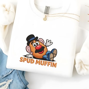May include: White sweatshirt with an embroidered cartoon potato character wearing a black hat and blue shoes. The text "SPUD MUFFIN" is in orange. A gold necklace and a clothing tag are also visible.