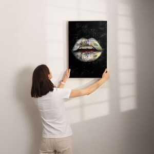 Puede incluir: Una impresi&oacute;n art&iacute;stica enmarcada que presenta un primer plano de labios, con los labios y los dientes hechos de billetes de cien d&oacute;lares estadounidenses. El fondo es negro. El arte se sostiene contra una pared blanca.