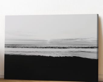 May include: Black and white canvas print of a beach scene. The bottom half of the image is a dark, black beach, while the top half shows the ocean and sky. The waves are white, and the sky is overcast.
