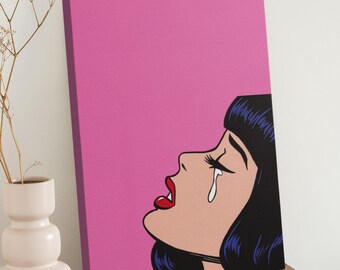 May include: Pop art canvas print featuring a crying woman with black hair, red lips, and a single tear. The background is a solid pink color. The artwork is displayed on a wooden surface.