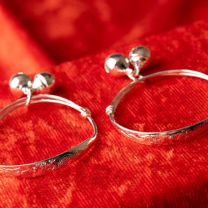 May include: Two silver baby bangles with small bells on top, resting on a red textured surface. The bangles have a delicate engraved design around the band. The bells are spherical and shiny.