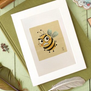 May include: A framed illustration of an angry cartoon bee with black and yellow stripes, set against a tan background. The bee has a furrowed brow and a stern expression. The artwork is in a white frame, resting on a green book.