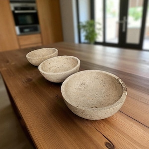 May include: Three textured, off-white bowls are arranged on a wooden table. The bowls have a speckled appearance and a rounded shape. The table has a natural wood grain finish, and the background is blurred.