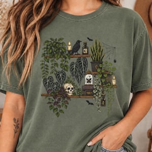 May include: An olive green t-shirt featuring a graphic print of wooden shelves holding various potted plants, a human skull, a black raven, lit candles, glass bottles, a butterfly in a glass dome, stacked books, a spider web, and small bats.