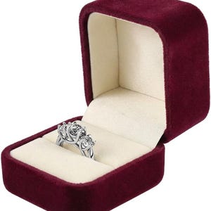 May include: A silver-toned ring featuring three round, faceted clear stones set in a prong setting, displayed inside an open, deep burgundy velvet jewelry box with a cream-colored interior lining and a hinged lid.