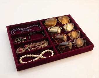 May include: A rectangular burgundy velvet jewelry organizer tray featuring multiple compartments. The tray holds four pairs of sunglasses with gold and brown frames, a purple beaded necklace, a tree pendant, a gold chain necklace, and a pearl necklace.