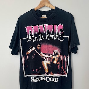 May include: Black t-shirt featuring the band name "DANZIG" in pink and white block letters above a graphic. The graphic depicts two figures and the text "SATANS CHILD" in white. The shirt is hanging on a wooden hanger.