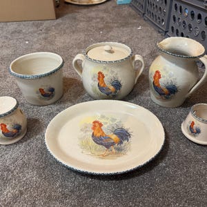 Puede incluir: Un juego de cocina de cerámica color crema con temática de gallo. El juego incluye una jarra, azucarero, plato y tazas. Cada pieza presenta una ilustración de gallo y ribete azul.