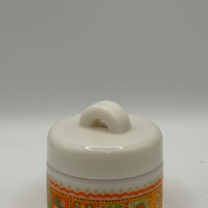 May include: A small, white ceramic jar with a matching lid and handle. The jar has a decorative band of orange, green, and yellow floral and geometric patterns around the base. The lid has a curved handle.
