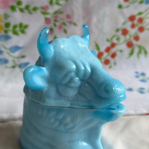 May include: A light blue milk glass lidded dish shaped like a cow head. The translucent glass features a molded design of a cow with horns, ears, and facial features. The lid sits atop a cylindrical base, creating a cohesive animal-shaped container.