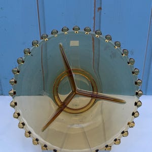 Könnte beinhalten: Ein Vintage-Servierteller aus bernsteinfarbenem Glas mit einem einzigartigen Design. Der Teller hat ein zentrales, dreizackiges Design und einen dekorativen Rand mit kleinen, bernsteinfarbenen Glasperlen. Der Teller wird auf einem transparenten Ständer vor blauem Hintergrund präsentiert.