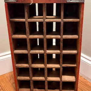 Puede incluir: Una caja de almacenamiento rectangular de madera con un marco exterior rojo y una rejilla interior de madera natural con veinte compartimentos individuales. La estructura incluye refuerzos metálicos en las esquinas superiores e inferiores.