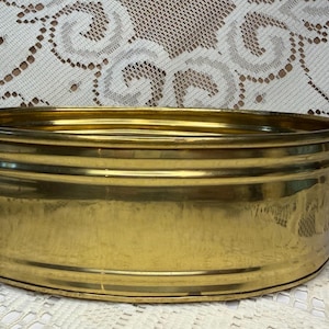 Pode incluir: Uma bacia oval de latão com acabamento metálico dourado polido, paredes arredondadas lisas com detalhes nervurados horizontais e duas pequenas alças integradas nas laterais. O recipiente tem um topo largo e aberto e uma base plana.