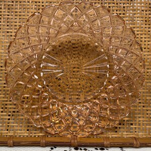 May include: A round, light pink glass plate featuring a textured diamond pattern around the outer rim and a central sunburst design with radiating lines. The glass is translucent, showing a geometric relief pattern across its entire surface.