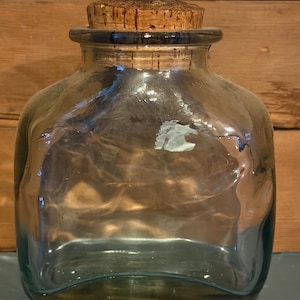 May include: A clear, square-shaped glass jar with a cork stopper. The jar has rounded edges and a wide opening. The cork is a light brown color. The jar is sitting on a blue surface, with a wooden background.