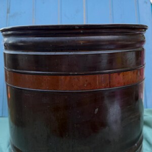Puede incluir: Un recipiente cilíndrico de color marrón oscuro con una banda de color cobre. El recipiente tiene una parte superior e inferior redondeadas, y la banda de cobre añade un acento de contraste. El recipiente parece ser de metal y tiene una estética vintage o antigua.