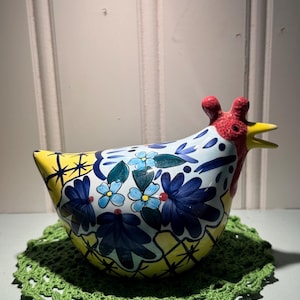 May include: A ceramic chicken figurine with a yellow body, blue floral designs, and a red comb. It rests on a green crocheted doily. The chicken's beak is yellow, and the background is a white wall.