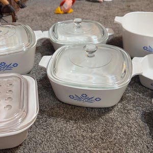 Puede incluir: Un conjunto de platos CorningWare blancos y azules con tapas de vidrio. Los platos presentan un diseño floral azul y están en varias formas y tamaños, incluyendo fuentes cuadradas y rectangulares y fuentes ovaladas.