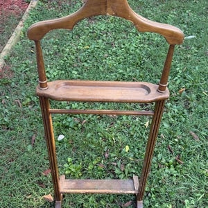 May include: A brown wooden valet stand featuring a wide contoured hanger top, a horizontal rod for trousers, a central recessed tray for accessories, and a lower shelf base. The structure is crafted from wood with visible wear and finish loss.