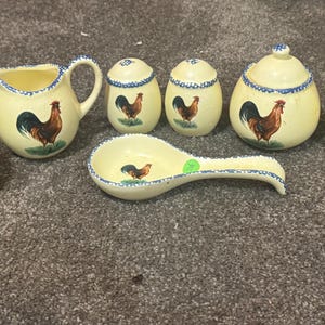 Puede incluir: Un conjunto de cerámica color crema para la cocina con un motivo de gallo. El juego incluye un azucarero, una jarra para crema, saleros y pimenteros, un reposacucharas, un tarro con tapa y un servilletero. Cada pieza está adornada con una detallada ilustración de gallo.