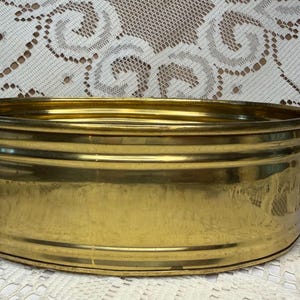 Puede incluir: Una palangana ovalada de latón con un acabado metálico dorado pulido, paredes redondeadas lisas con detalles acanalados horizontales y dos pequeñas asas integradas en los laterales. El recipiente tiene una parte superior abierta y base plana.