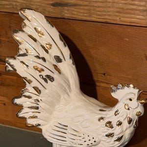 Puede incluir: Figura de gallo de cerámica blanca con detalles dorados. El gallo tiene plumas detalladas y una base decorativa. Los detalles dorados se aplican a las plumas de la cola, la cresta y la base. La figura está sobre un fondo de madera.