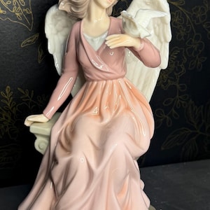 May include: A porcelain angel figurine in a seated pose, wearing a flowing pink dress and a matching wrap. The angel has white wings and holds a white dove. The figurine is on a base with flowers and is set against a dark background.