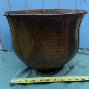Può includere: Un grande vaso in metallo color bronzo antico con un bordo svasato e una superficie martellata e strutturata. Il vaso misura circa 25 cm di diametro. Lo sfondo comprende una staccionata di legno blu e un panno verde.