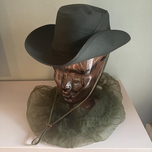 May include: An olive green hat with a wide brim and a round crown. The hat has a chin strap and is displayed on a mannequin head. A green mesh net is draped around the mannequin's neck.