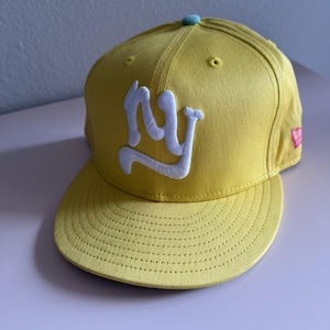 May include: Yellow baseball cap with a curved brim. The cap features a white embroidered "NY" logo on the front. The top button is light blue, and a pink logo is visible on the side.