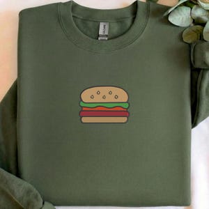 May include: Olive green sweatshirt featuring a cartoon burger design. The burger has a bun, lettuce, tomato, and a patty. The design is centered on the front of the long-sleeved shirt. The shirt is made of a soft material.