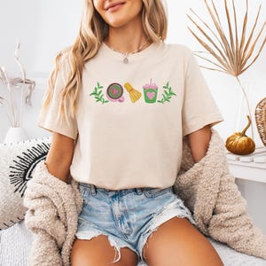 May include: Beige t-shirt with a matcha-themed graphic. The design includes a matcha bowl, whisk, and drink, all in green, pink, and yellow. The shirt is worn by a person with blonde hair and blue jean shorts.