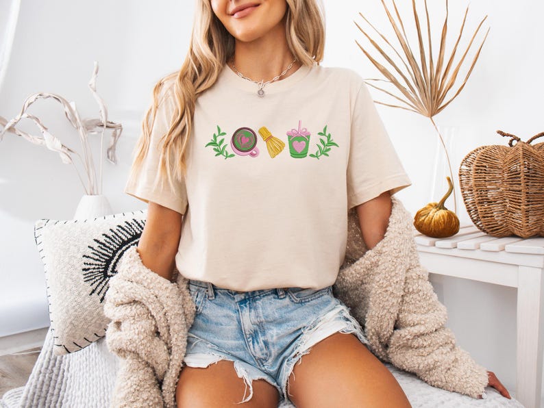 May include: Beige t-shirt with a matcha-themed graphic. The design includes a matcha bowl, whisk, and drink, all in green, pink, and yellow. The shirt is worn by a person with blonde hair and blue jean shorts.