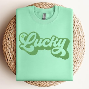 May include: A light green sweatshirt with the word "Lucky" embroidered in a darker green, stylized font. The design includes a small shamrock. The sweatshirt is displayed on a round, woven, light brown placemat.