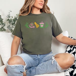 May include: Olive green t-shirt with embroidered matcha-themed designs. The shirt features a matcha bowl, whisk, and drink with a heart. The model is wearing ripped blue jeans.