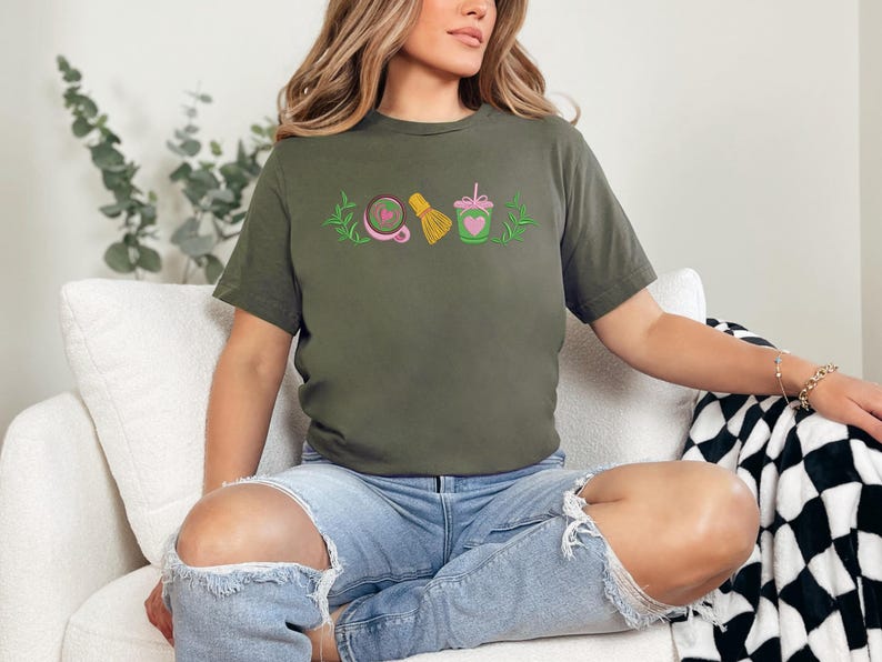 May include: Olive green t-shirt with embroidered matcha-themed designs. The shirt features a matcha bowl, whisk, and drink with a heart. The model is wearing ripped blue jeans.