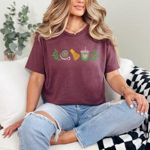 May include: A maroon t-shirt featuring an embroidered design of matcha tea elements. The design includes a whisk, a bowl, a heart-shaped matcha drink, and a sprig of leaves. The shirt is worn by a person wearing ripped blue jeans.