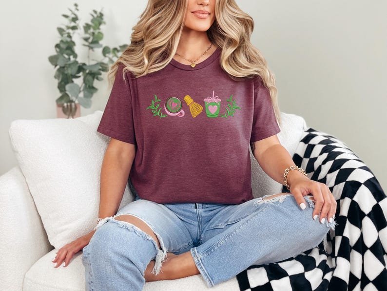 May include: A maroon t-shirt featuring an embroidered design of matcha tea elements. The design includes a whisk, a bowl, a heart-shaped matcha drink, and a sprig of leaves. The shirt is worn by a person wearing ripped blue jeans.