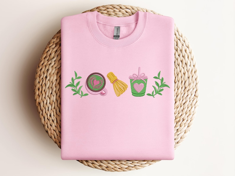May include: A light pink sweatshirt featuring embroidered matcha-themed designs. The design includes a matcha latte with a heart, a bamboo whisk, and a matcha drink with a heart. Green leaves are on either side of the design.
