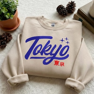 May include: A beige sweatshirt with the word "Tokyo" in blue script and Japanese characters. The sweatshirt has a crew neck and cuffed sleeves. The design includes four blue stars above the word "Tokyo".