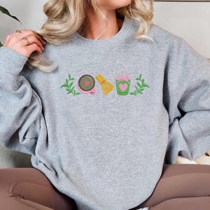 May include: A light gray sweatshirt with embroidered matcha-themed graphics. The design includes a pink teacup with a heart, a yellow whisk, and a green cup with a heart and straw, all framed by green leaves.
