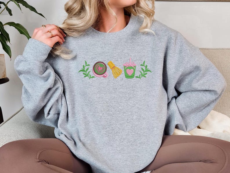 May include: A light gray sweatshirt with embroidered matcha-themed graphics. The design includes a pink teacup with a heart, a yellow whisk, and a green cup with a heart and straw, all framed by green leaves.