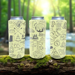 May include: Three insulated beverage holders with silver tops and a light yellow background. Each holder features a different black and gray outdoor-themed design, including mountains, trees, and camping gear. The word "ADVEN" is visible on one.