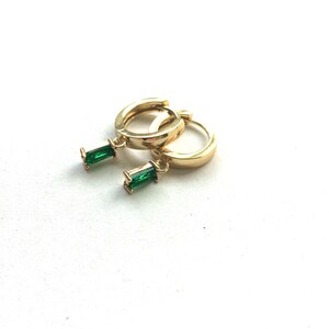 Könnte beinhalten: Ein Paar goldfarbene Creolen-Ohrringe mit jeweils einem kleinen, rechteckigen, smaragdgrünen Edelstein. Die Ohrringe sind vor einem weißen Hintergrund abgebildet.