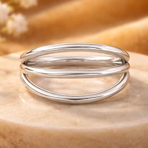 Op de afbeelding: Een gepolijste zilverkleurige ring met een ontwerp van drie banden, bestaande uit drie gladde, afgeronde, parallelle metalen lussen die aan de zijkanten zijn verbonden tot één samenhangend sieraad met een reflecterende afwerking.