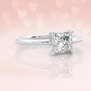 May include: A silver ring featuring a square-cut diamond. The ring is set against a light pink backdrop with blurred white heart shapes. The diamond sparkles, reflecting light. The ring's band is slender and polished.