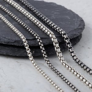 Puede incluir: Cuatro collares de cadena de caja plateados de diferentes tonos se muestran sobre una superficie texturizada gris oscuro. Las cadenas, hechas de pequeños eslabones rectangulares, varían de plata brillante a un acabado más oscuro y oxidado.